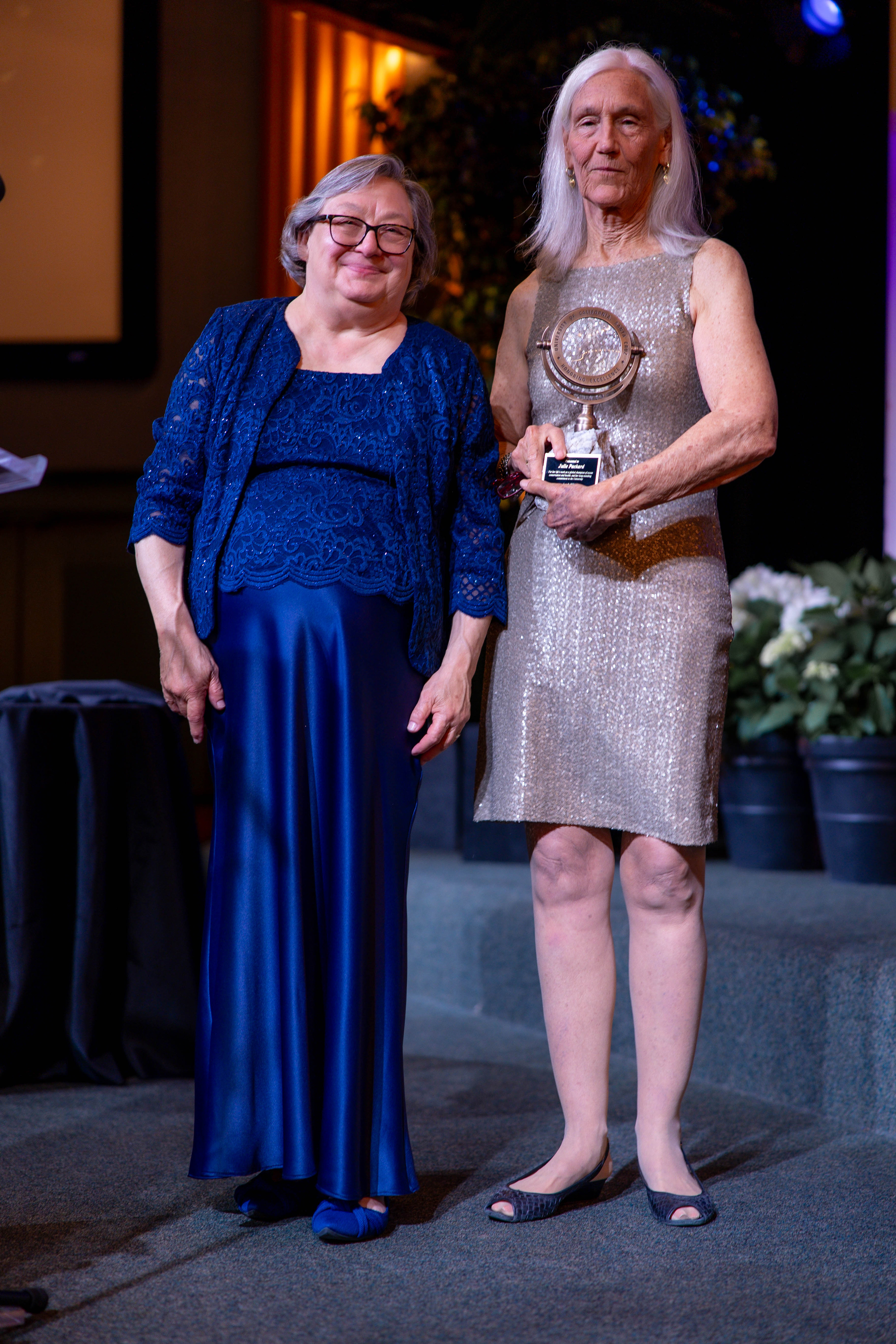 UC Santa Cruz Chancellor Cynthia Larive, in a blue gown, left, next to Julie Packard, in a silvery gown, right. Packard is holding an award Chancellor Larive has given her.
