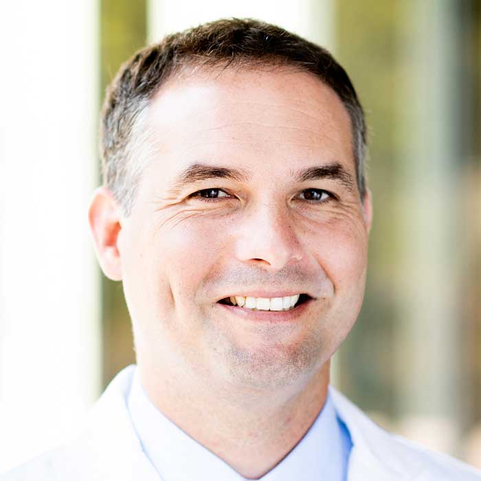 Man with short dark hair wearing a white coat smiles at the camera