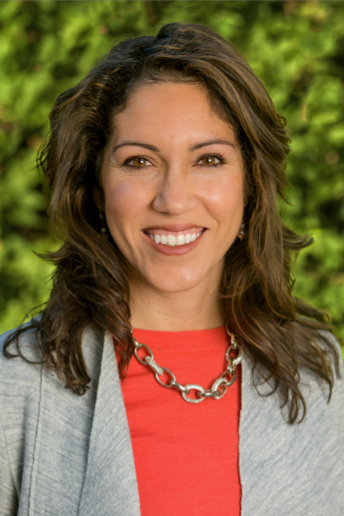 Maria Anguiano, woman with wavy shoulder-length hair in a gray jacket and red top with a necklace, smiling