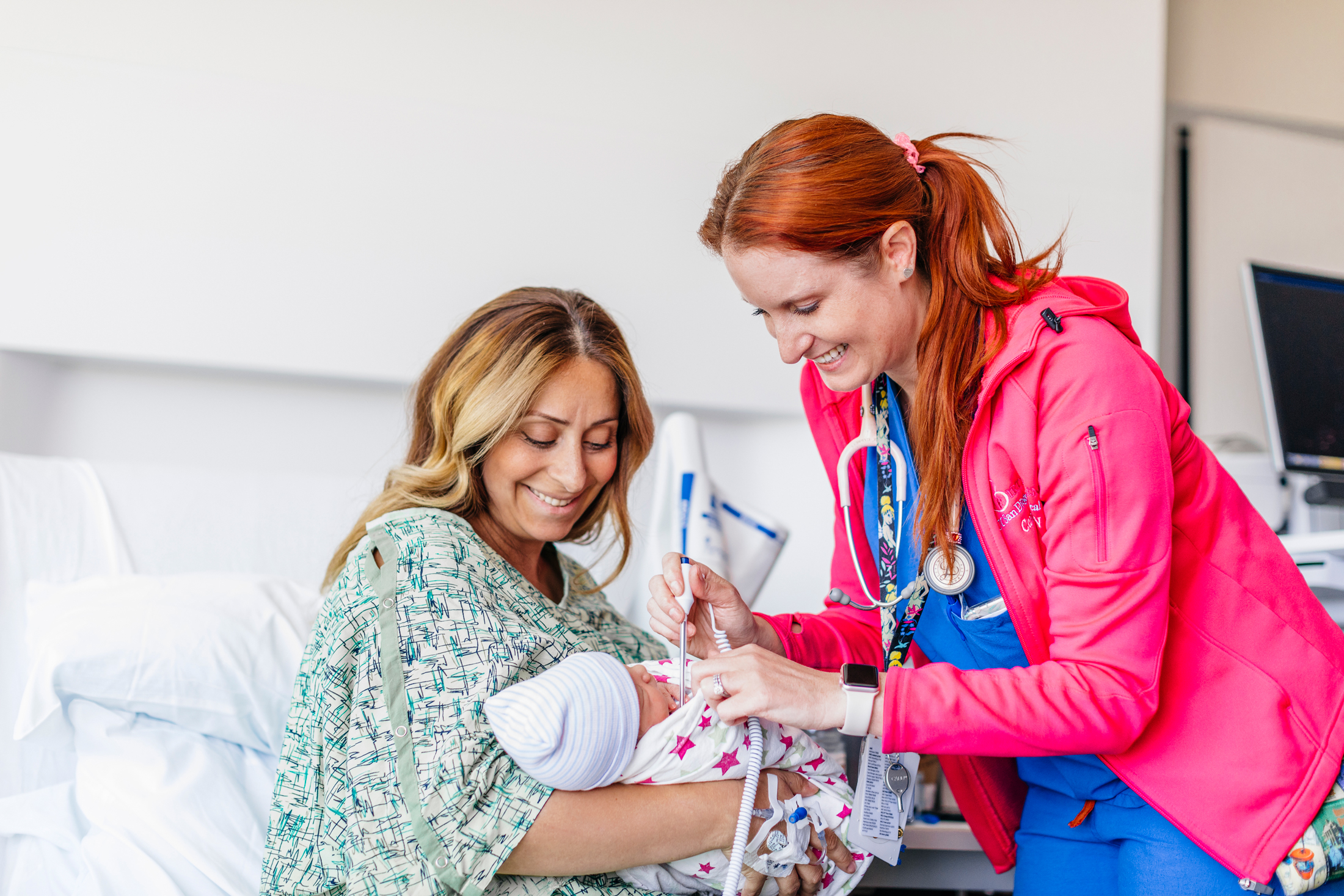 A smiling nursing mother holds her baby in the hospital while a nurse with red hair helps her