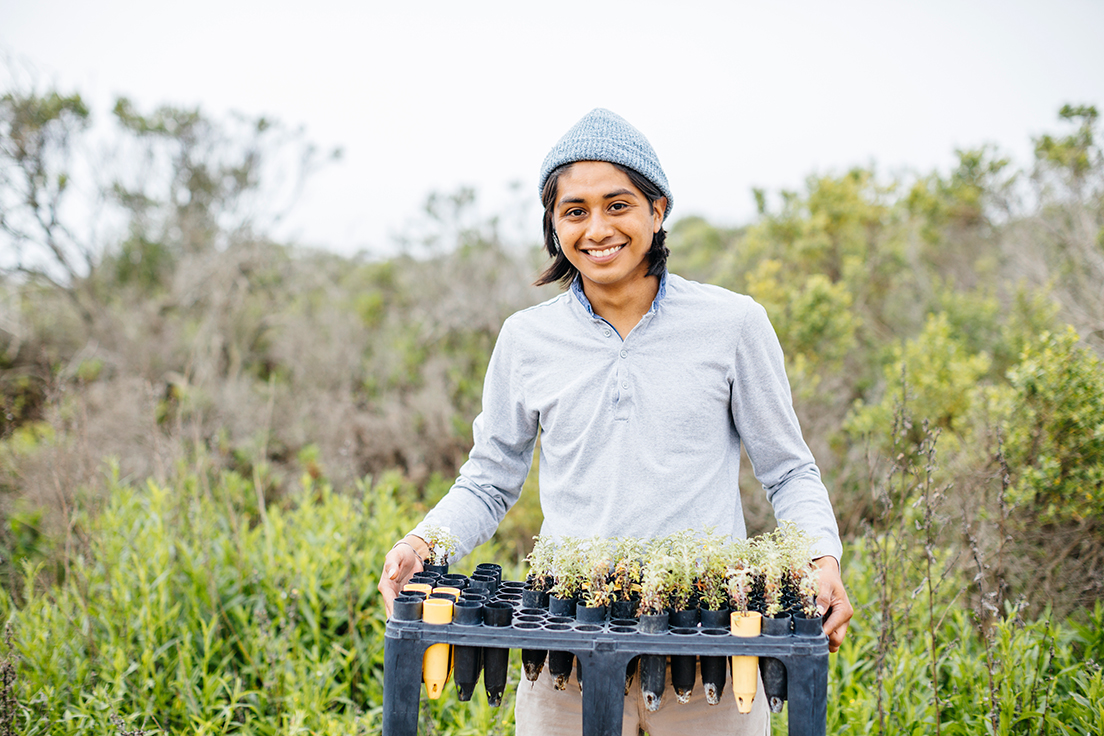 A student with a beanie holds up a tray of seedlings while in a green and wild environment
