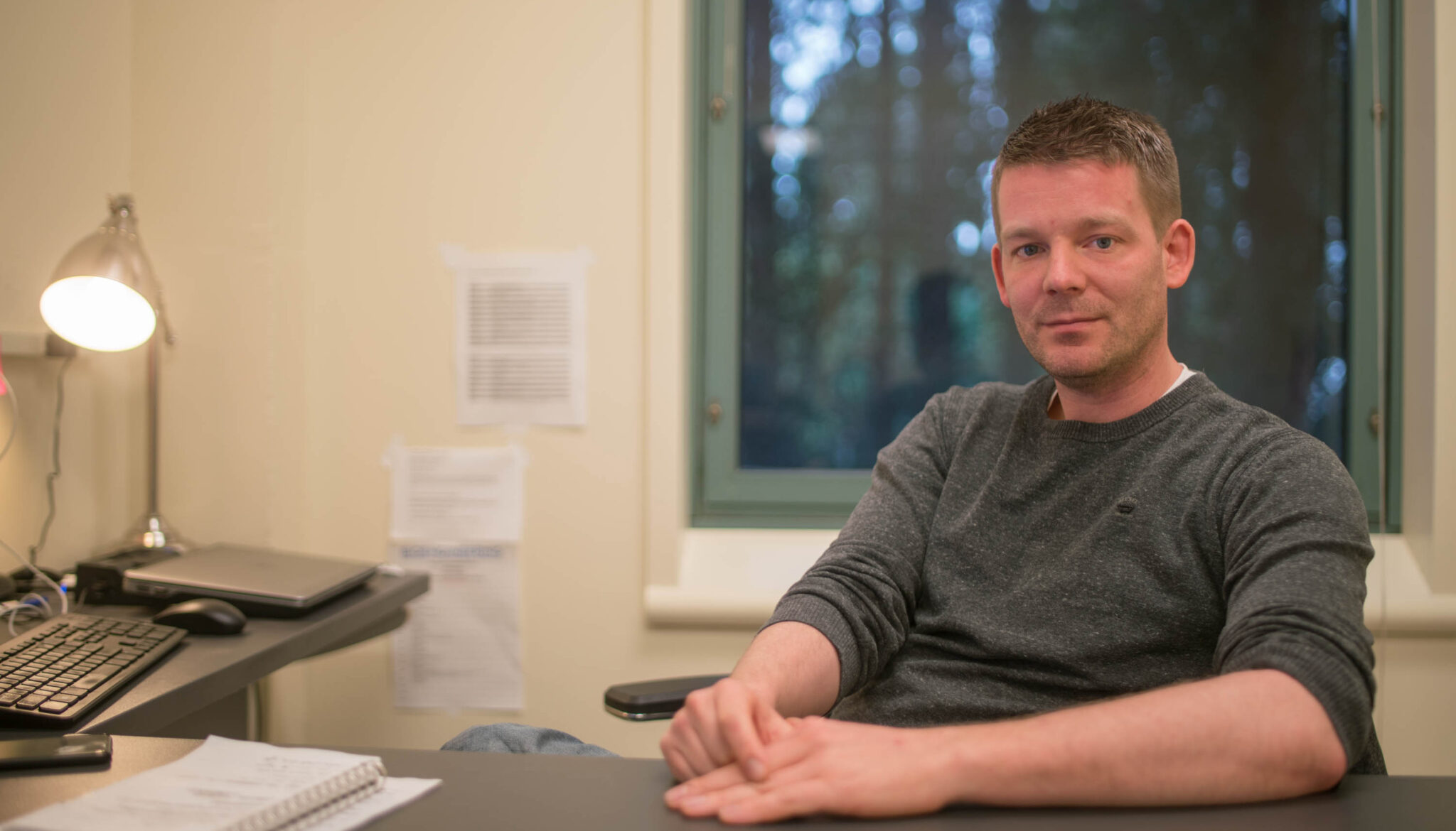 Man in long-sleeved shirt sits at desk with keyboard and a window showing the evening and trees behind
