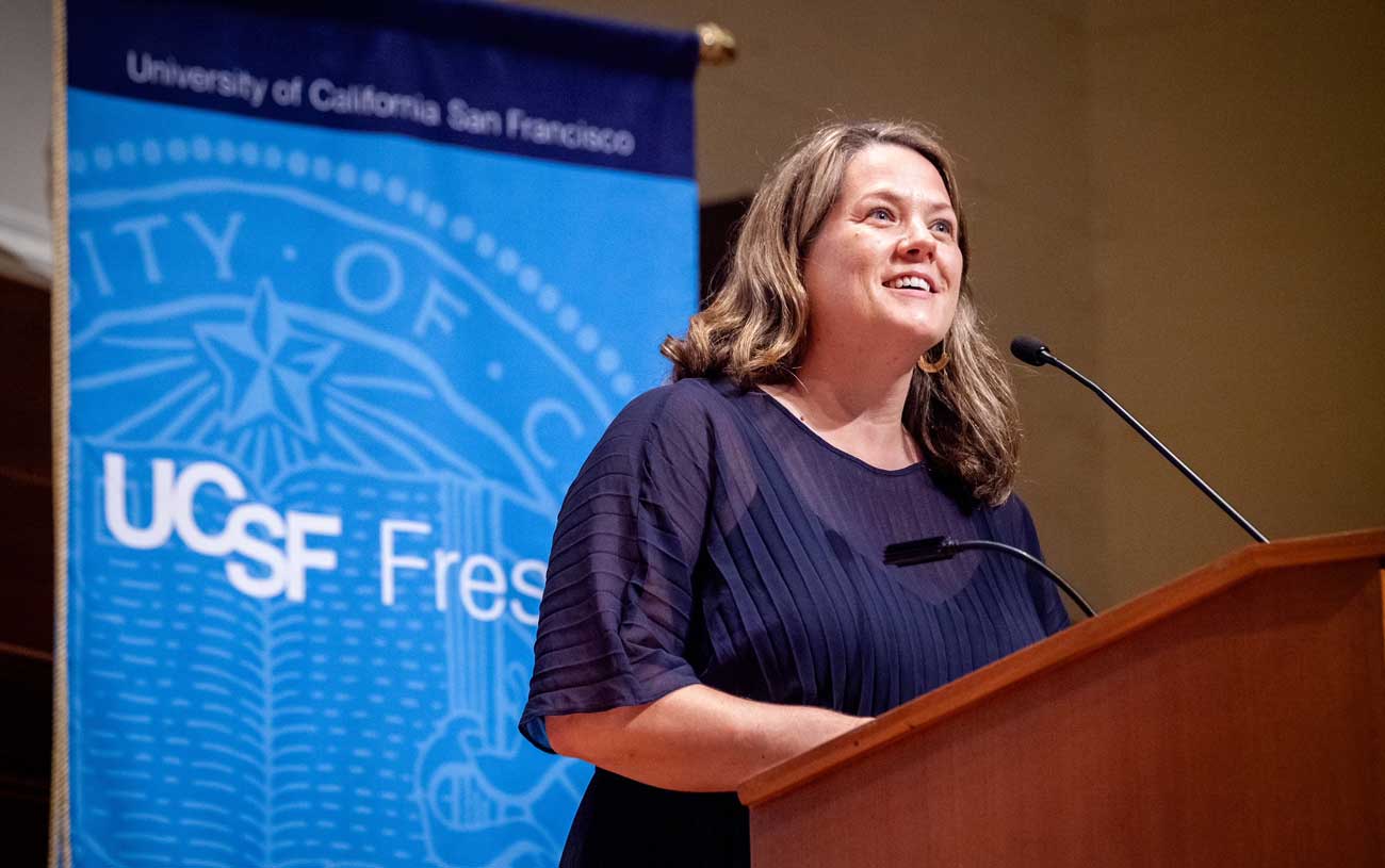 Woman with shoulder-length blonde hair at a lectern with a UCSF Fresno sign behind