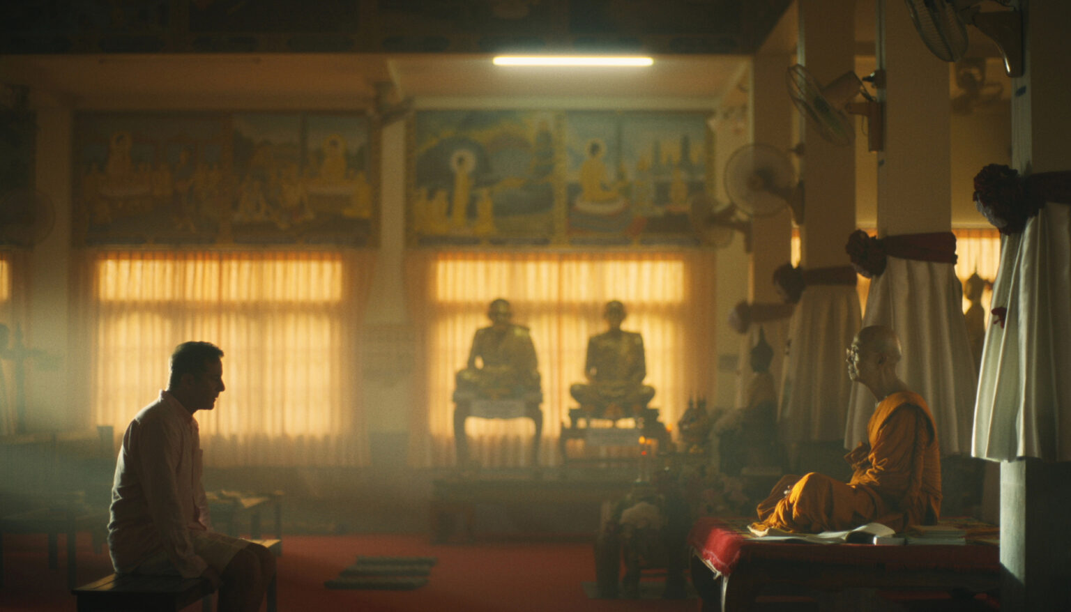The character of Tim Ratliff kneels in front of a monk, both in profile, in a monastery, as two other monks look on