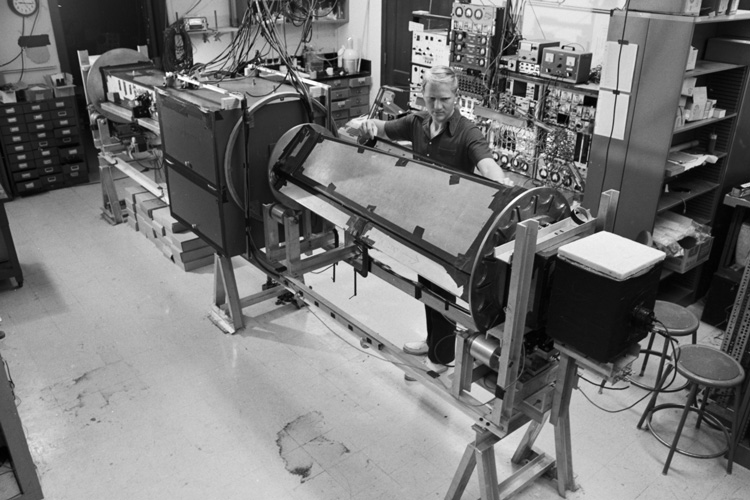 John Clauser stands next to a giant, retro-looking machine in a lab in a black and white photo from the 1970s.