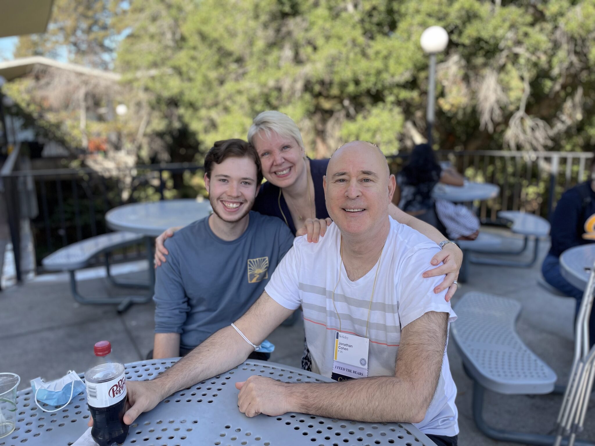 A young man smiles at a plastic table with his parents. The mother has her arms around the father and son, who are seated. There is a blue mask on the table and on the father's arm, and the dad wears a name tag