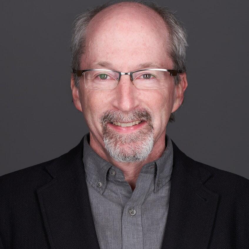 Man with glasses and a beard in a dark suit jacket and gray dress shirt smiles at the camera