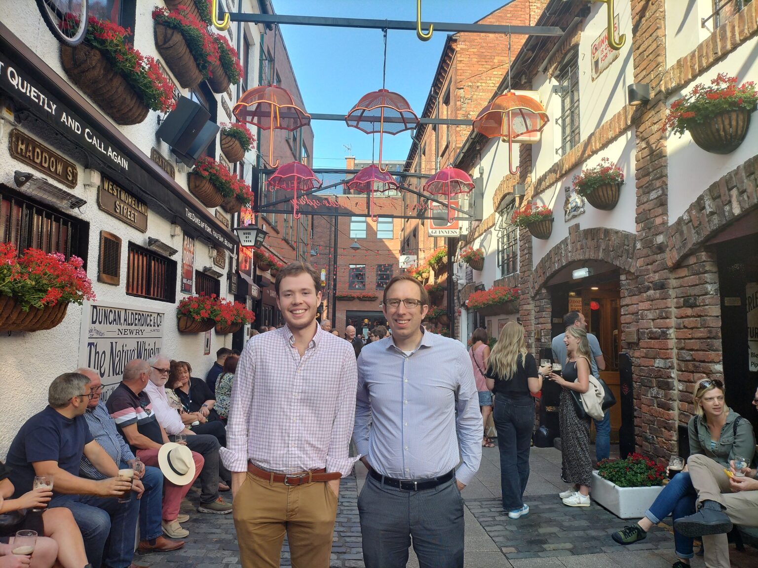 two men in collared shirts stand in a street with whimsical umbrella decorations overhead