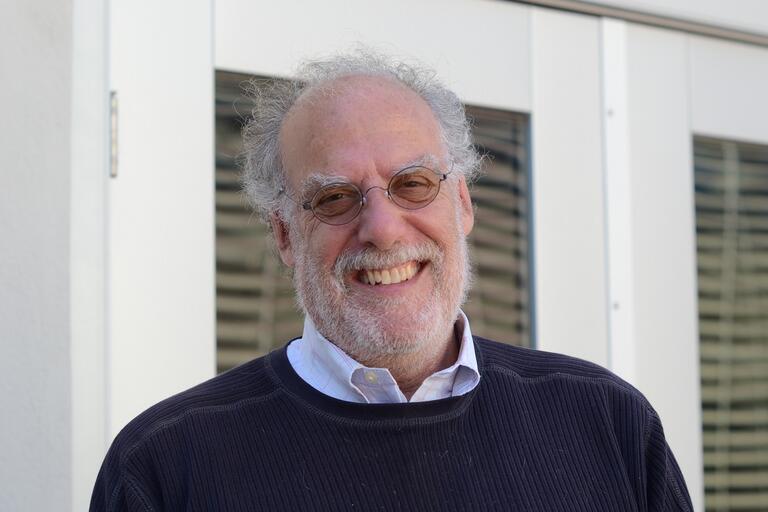 Steven Kahn smiles at the camera wearing glasses, a sweater and button-up shirt.
