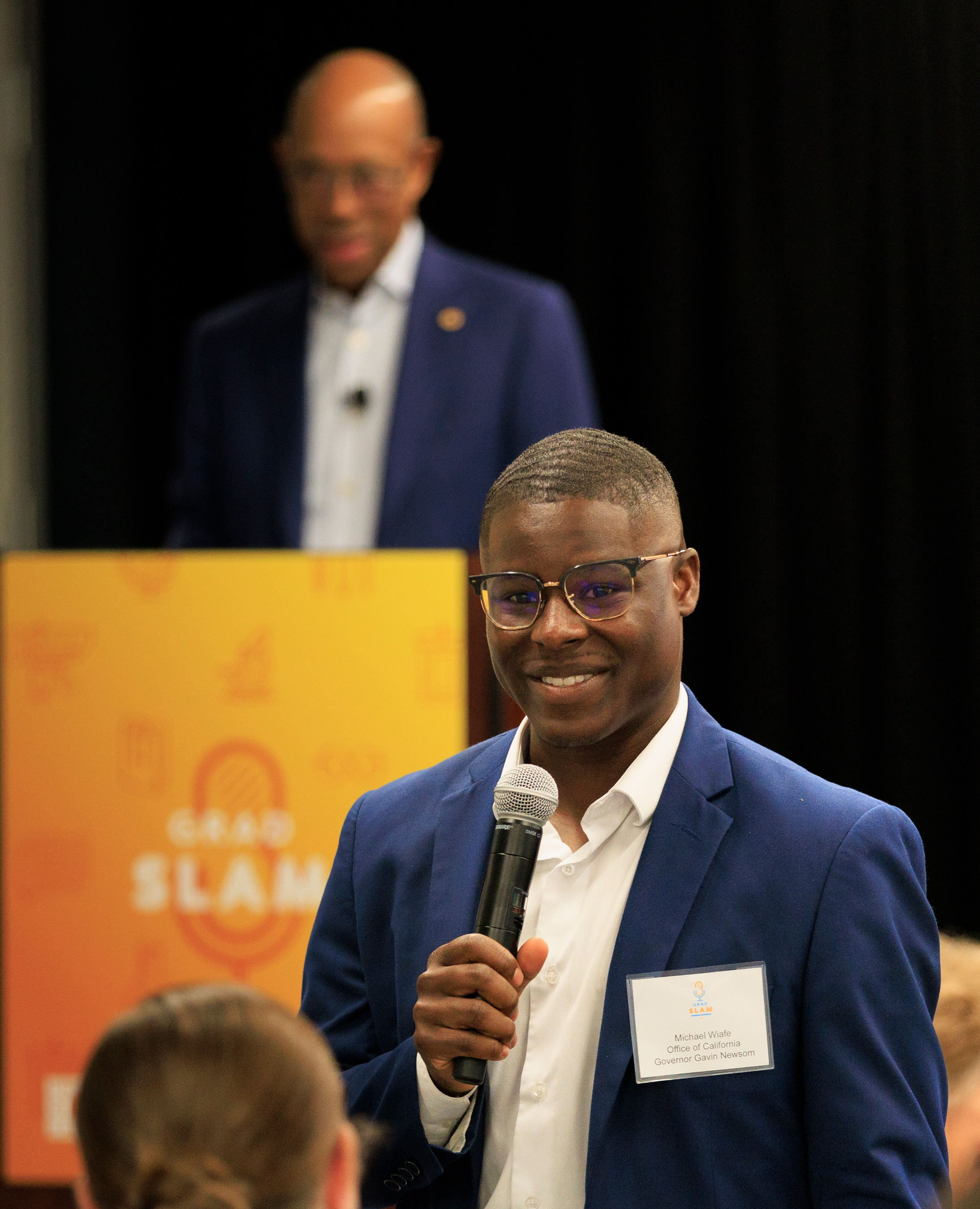 Young man in a blue suit jacket speaks into a mic with President Drake behind him out of focus at a Grad Slam lectern