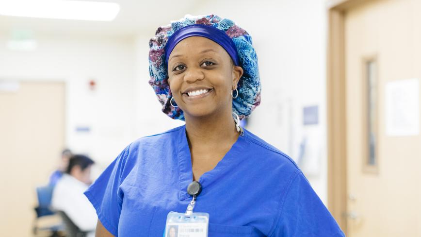 A nurse in purple scrubs and cap smiles at the camera