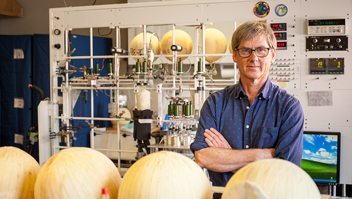 Ralph Keeling poses for a portrait with his arms crossed, standing in front of a complicated wall of instruments in a lab