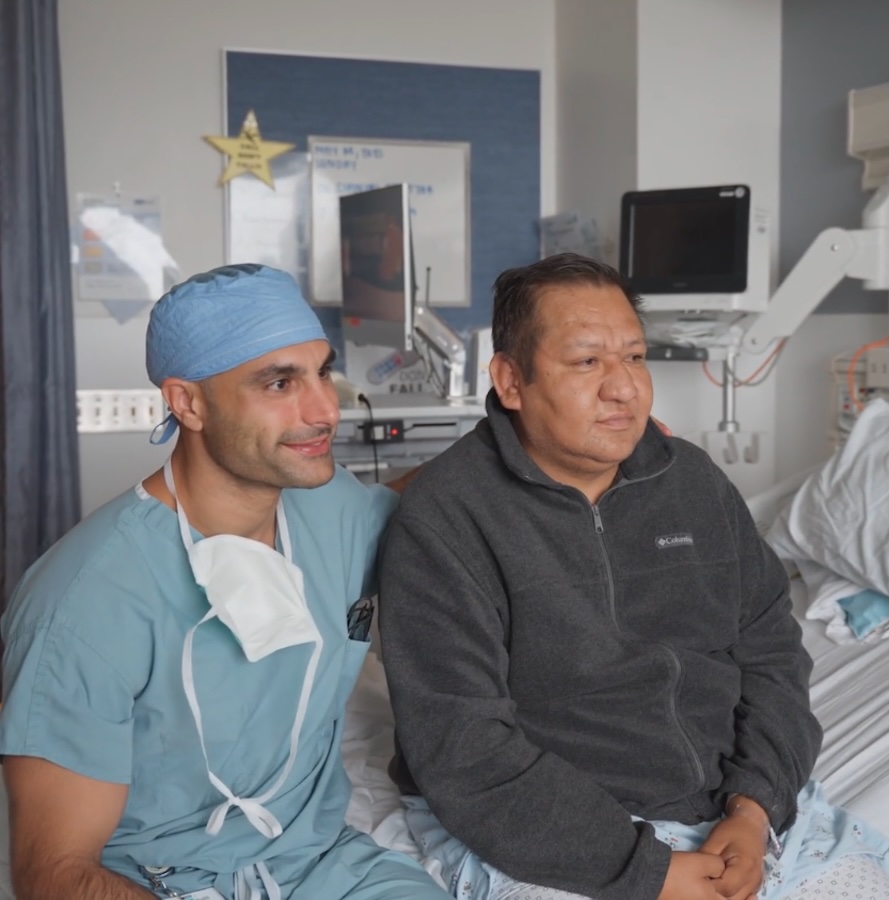 A surgeon in scrubs and a patient sitting up in a hospital bed pose together