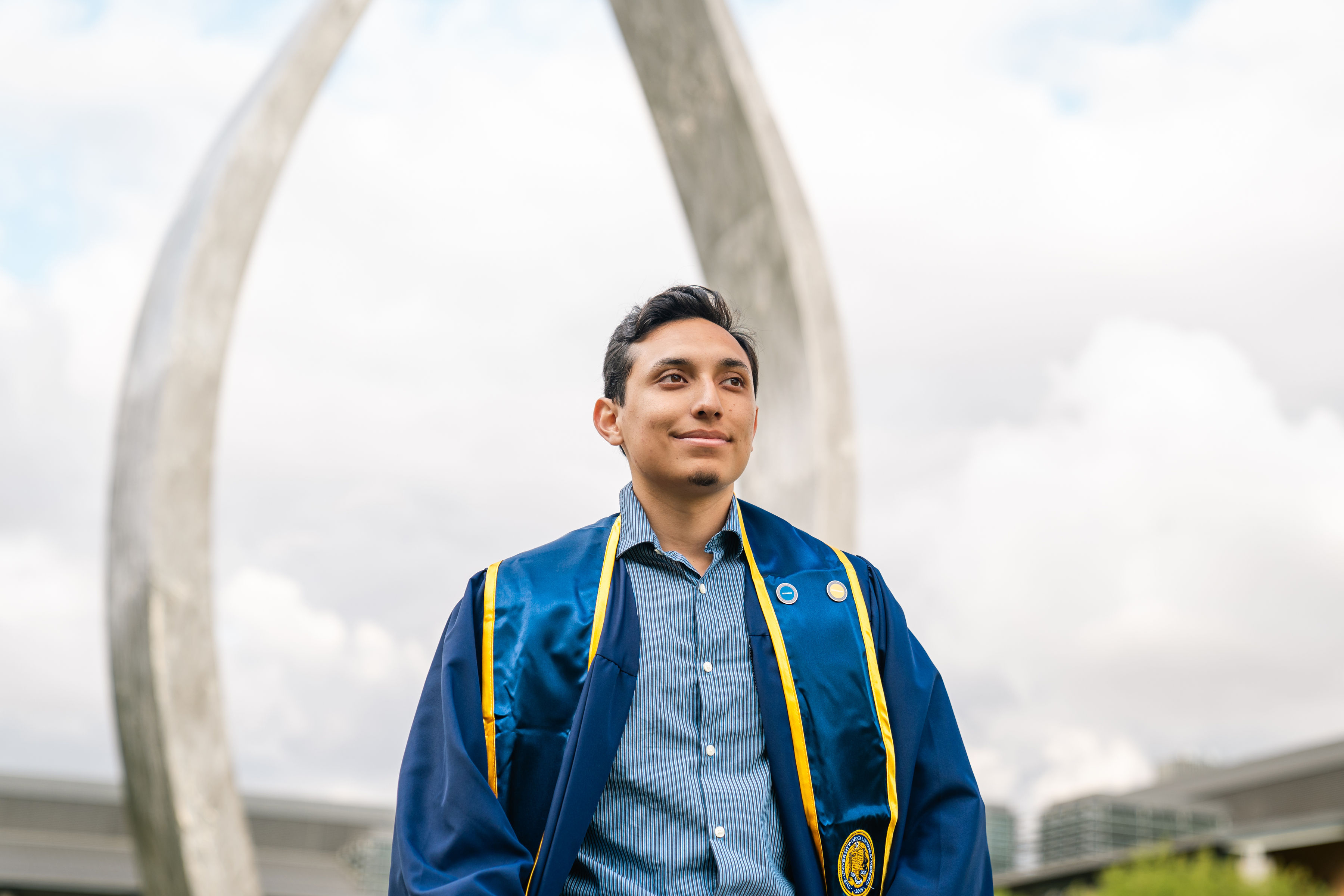 A young man in a stole and blue graduation robe in front of the UC Merced Beginnings sculpture with two rising metal arms