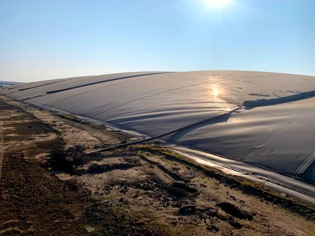 A dark tarp covers a large amount of dry farmland, sun shining brightly on the top edge of the photo