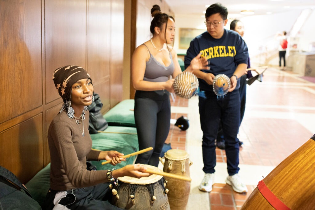Three students play drums and various other percussion instruments in a hallway
