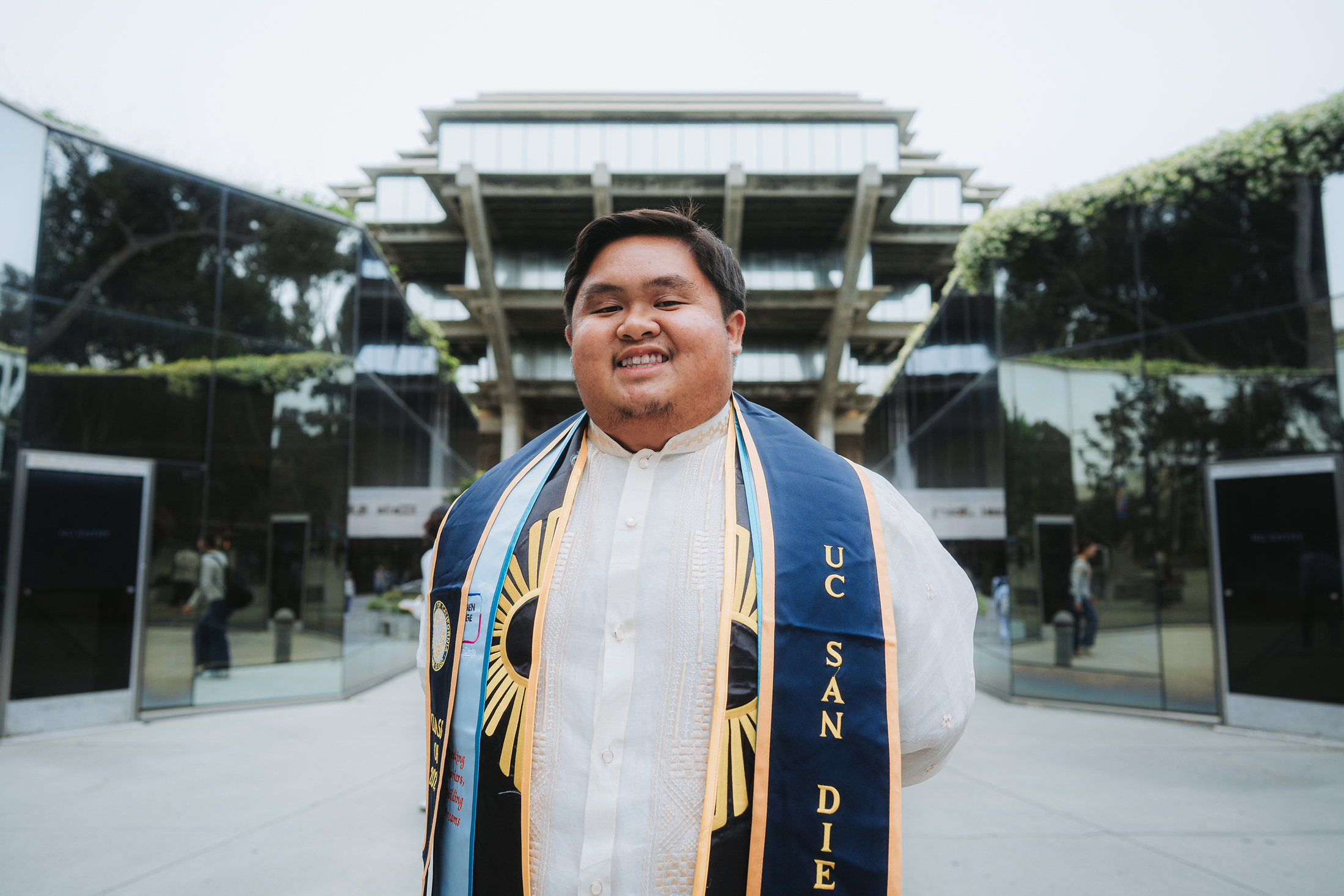 A young man with graduation stole smiles in front of UC San Diego's Geisel Library