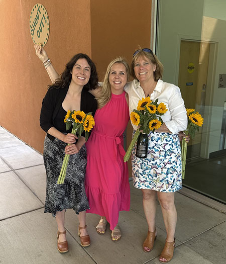 Three women pose together, holding sunflower bouquets