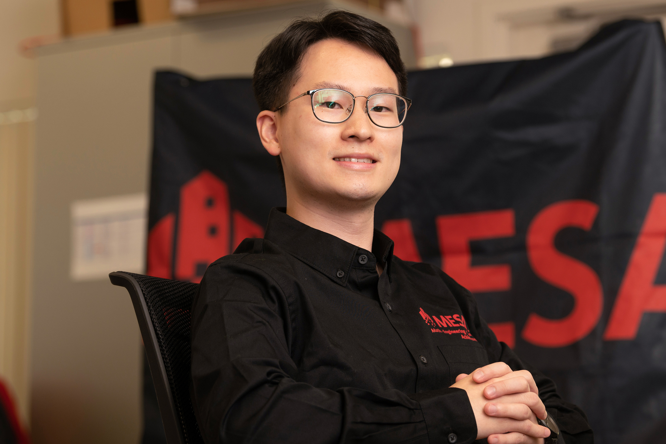 Young man with glasses sits in a chair with hands folded together, MESA flag behind