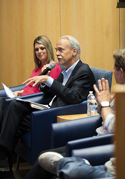 In a wood-paneled room, a seated man, center, gestures as he talks into a microphone. A woman looks on in the chair to his right