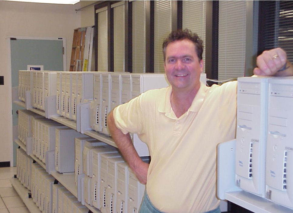 A smiling man in a yellow polo shirt standing by bank of computers