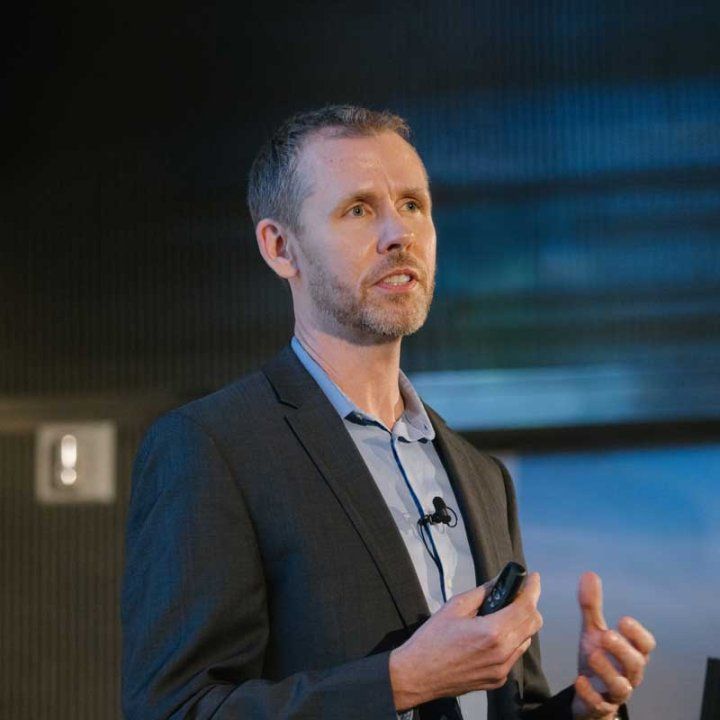 A man with a mic on his dress shirt gestures with his hands while giving a lecture