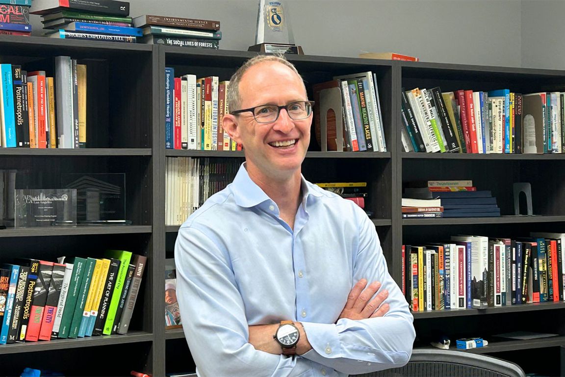 A man with glasses and his arms crossed smiles in front of bookshelves in an office