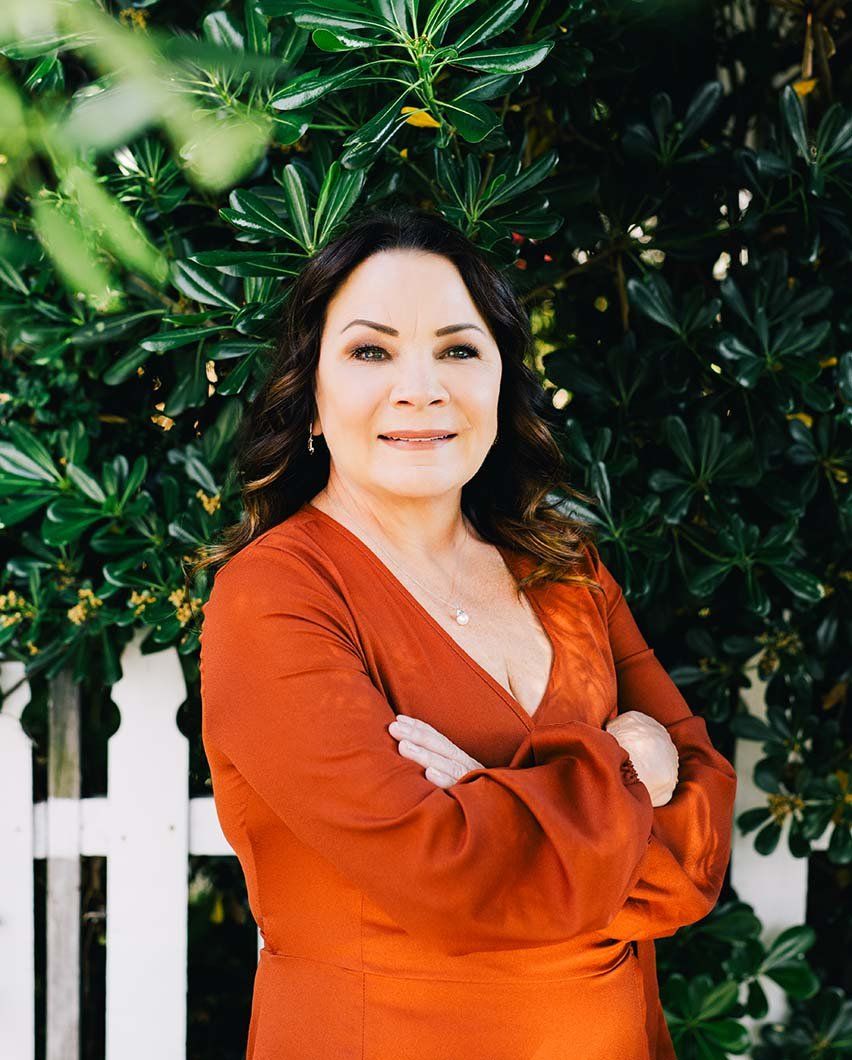 Woman in orange dress with arms crossed and long black hair poses outside, smiling
