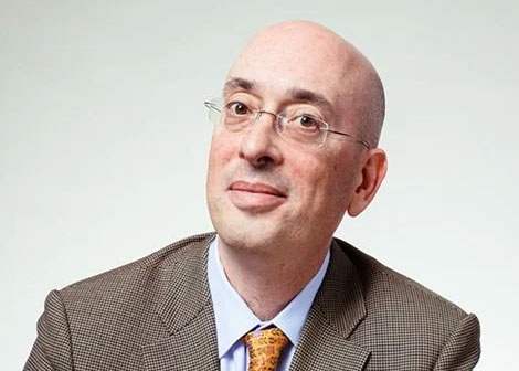 Adam Boxer smiles at the camera, wearing a tweed jacket and yellow tie. 