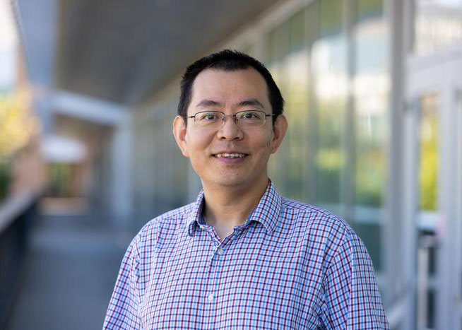 Jinyong Liu, wearing a plaid shirt, smiles at the camera standing in front of a bank of plate glass windows.