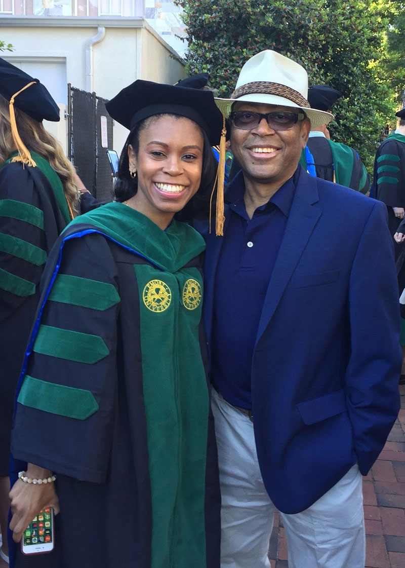 Mel Mann and Patrice Mann, wearing graduation attire, smile for a photo with their arms around each other's shoulders
