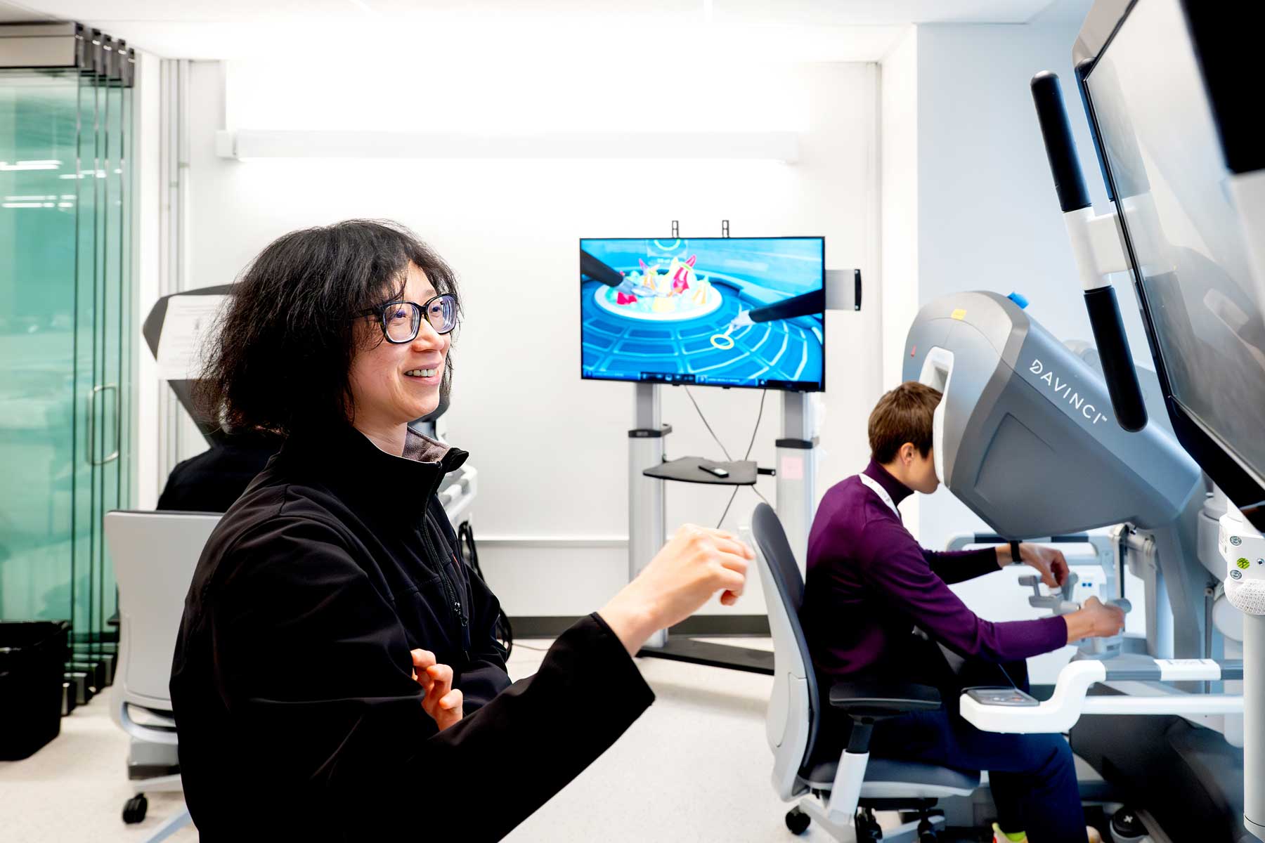 A doctor in an exam setting looks at a screen on the wall, while a person in the background sits at a desk with their face pressed against a large machine for viewing images. 
