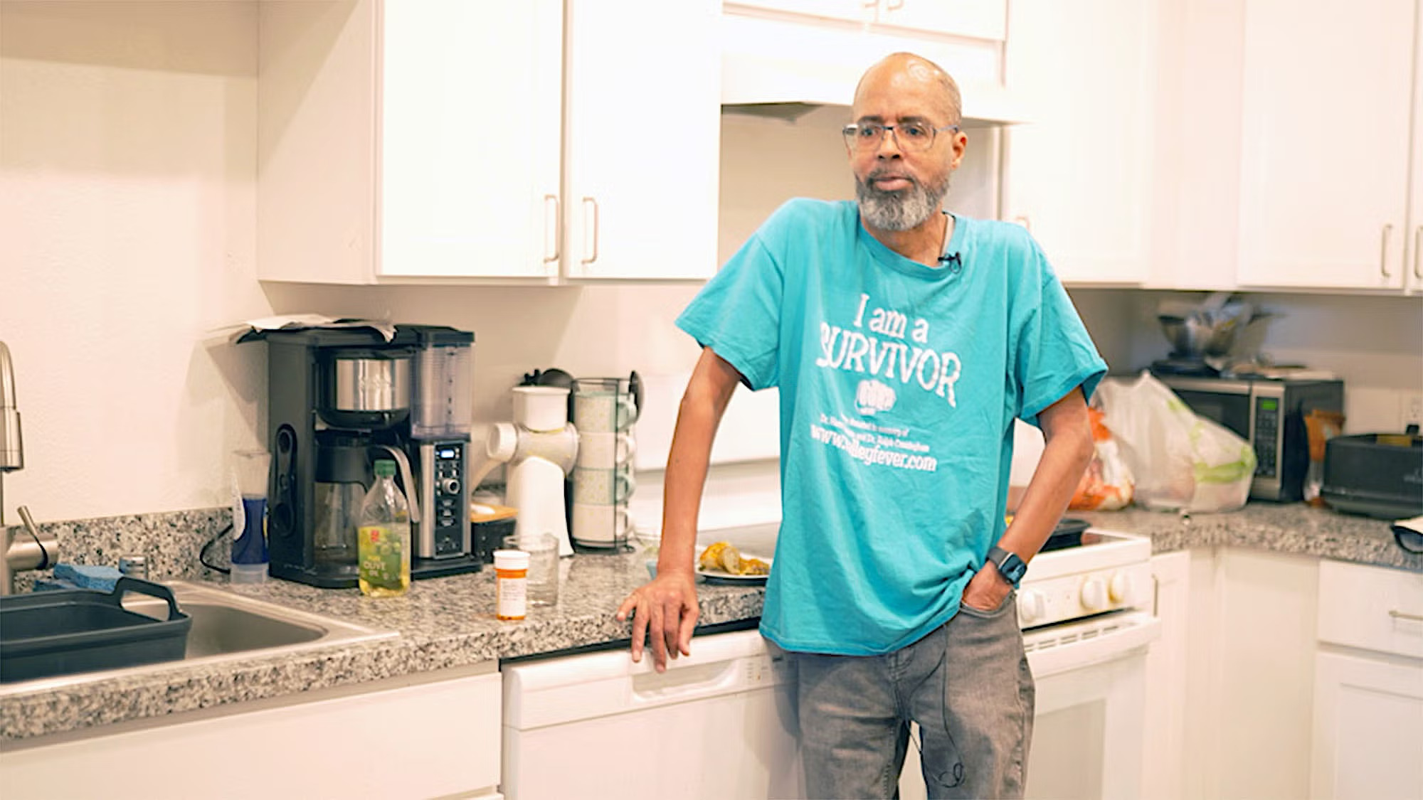 A man with a goatee and a shirt that says "I Am A Survivor" leans against a kitchen counter