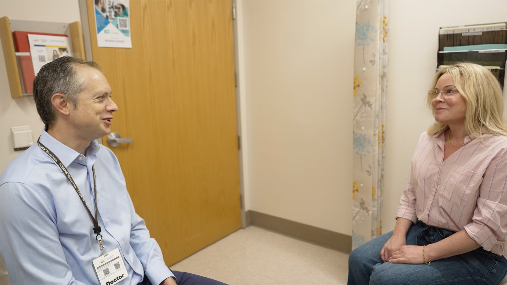 A man on the left with a badge talks to a woman in an exam room, both seem relaxed and happy