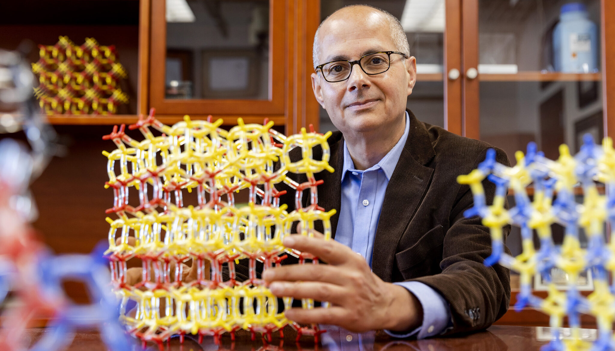 A man with glasses and a brown jacket holds molecular models on a table