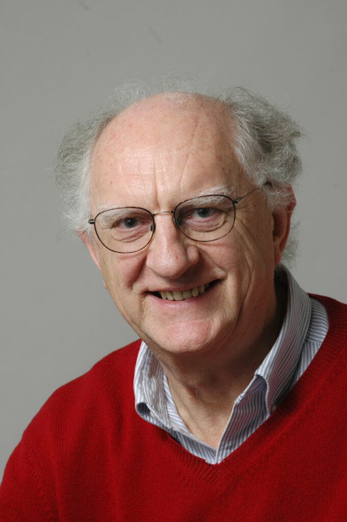 John Clarke smiles at the camera wearing a red sweater.