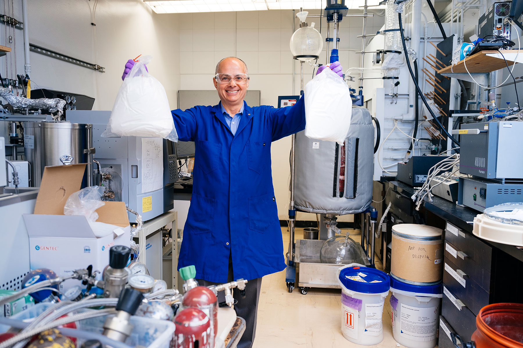 A man with glasses in a blue lab coat holds up two bags in the lab