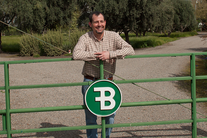 A man leans against a farm gate with a B on it