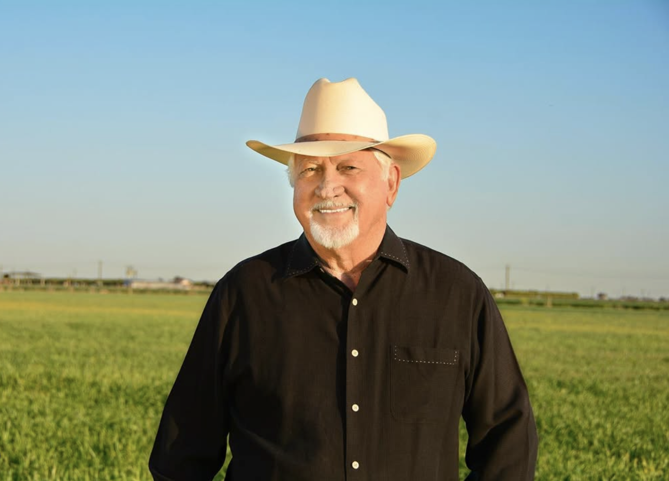 A man in a white cowboy hat with a white goatee stands in front of a field smiling