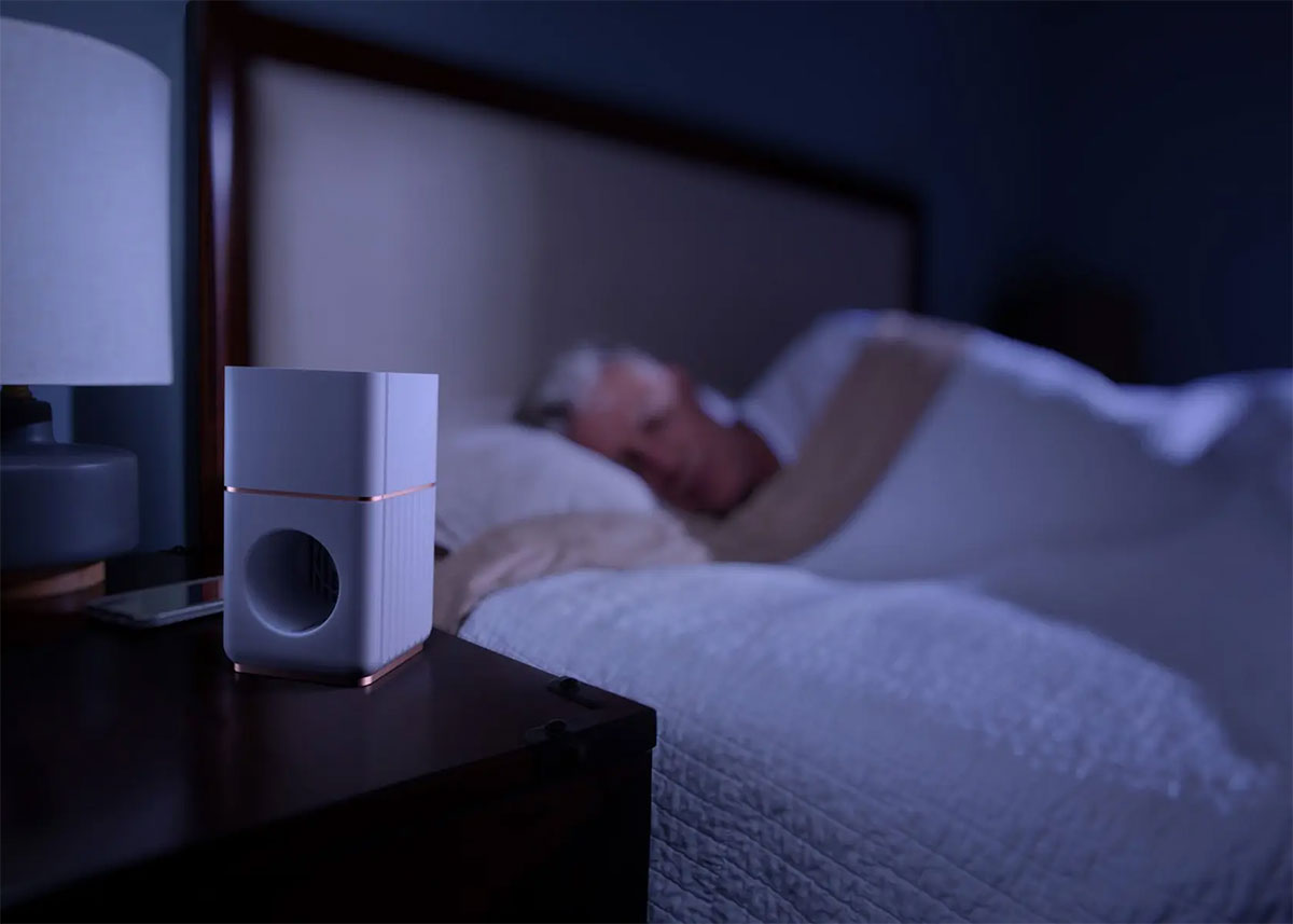 A white device about the size of a coffee mug sits on a bedside table next to an older gentleman asleep in bed