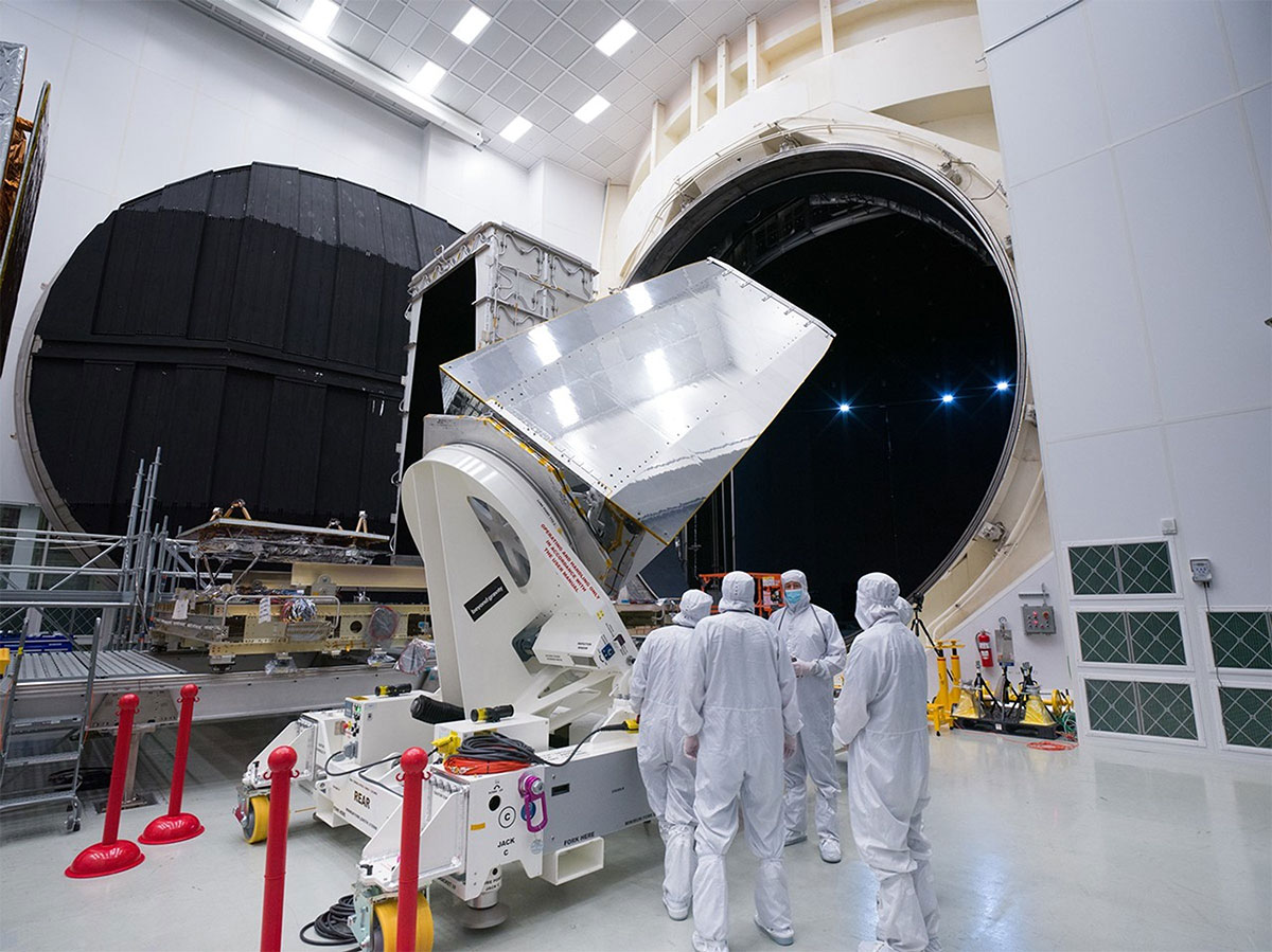 Five people in white sterile suits stand next to a large telescope mirror being loaded into a huge testing chamber in a sterile room