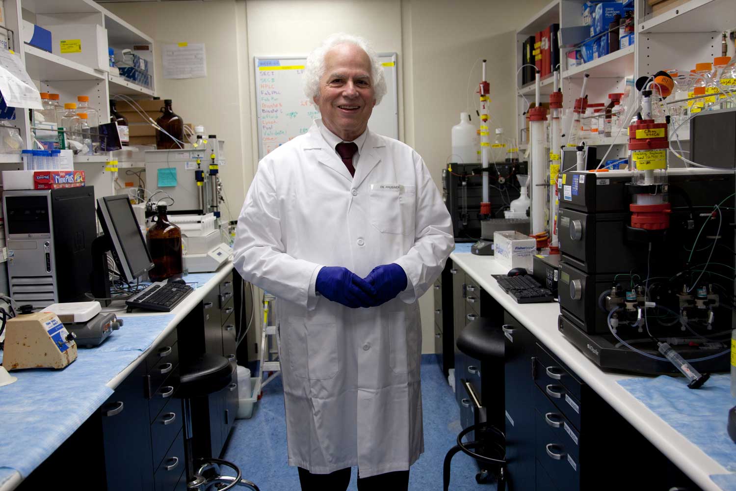 A man in a lab coat and blue medical gloves smiles in a lab