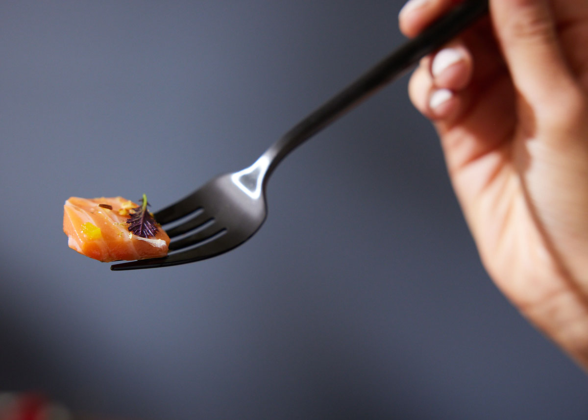 A hand holds a fork with a bite of salmon on it, against a gray background