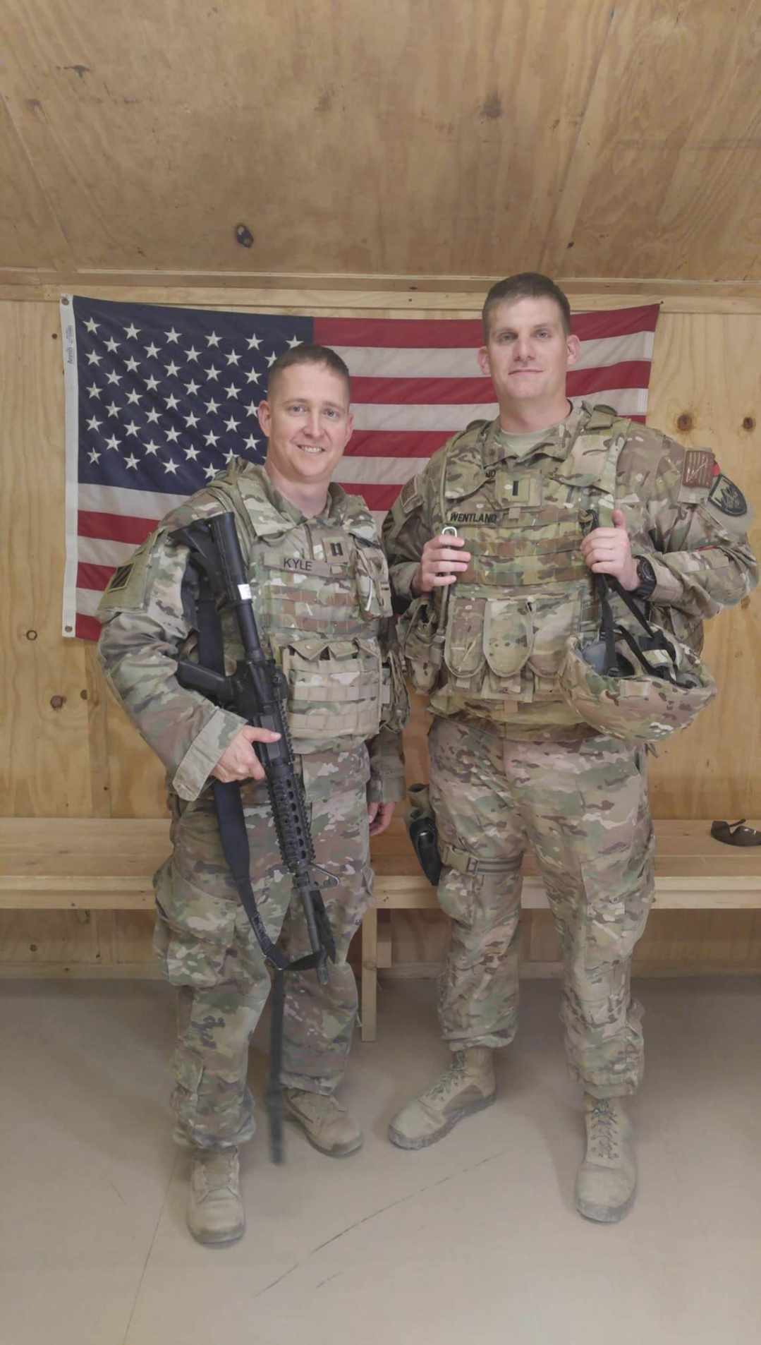 Two soldiers in uniforms and flak jackets, one holding a firearm, posing before an American flag