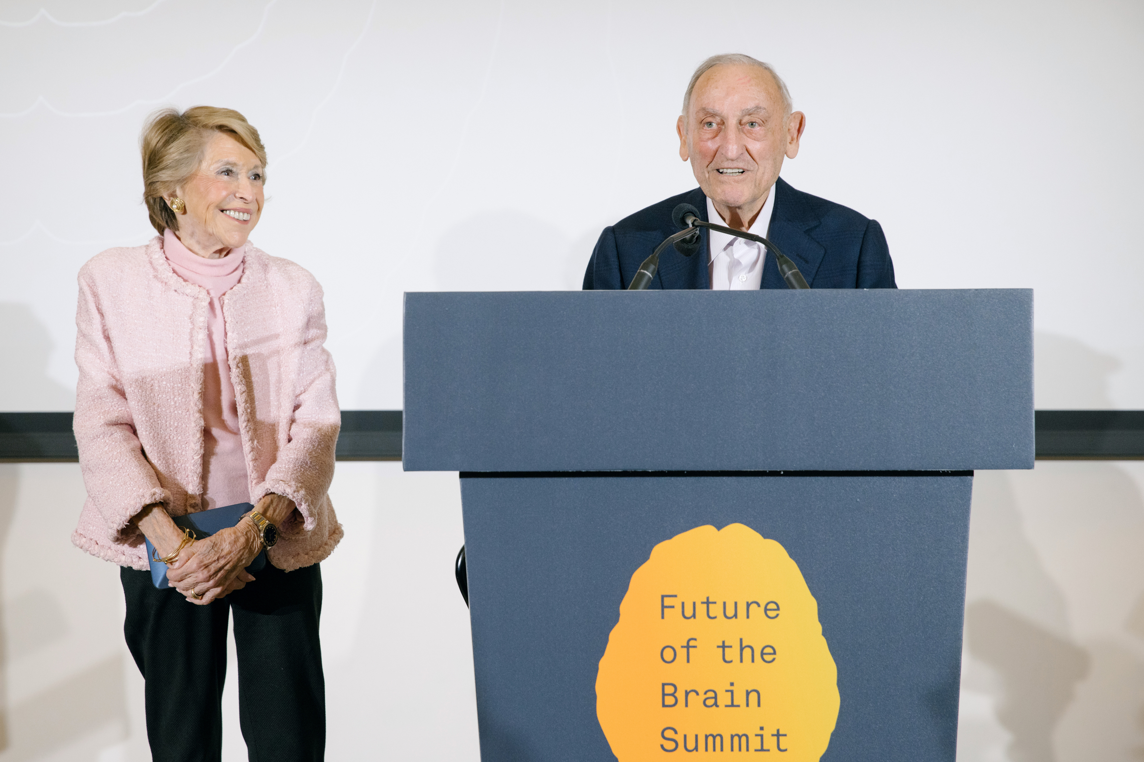 Joan Weill in a pink jacket and blue stands and smiles while looking at Sandy Weill, at a blue podium that says Future of the Brain Summit
