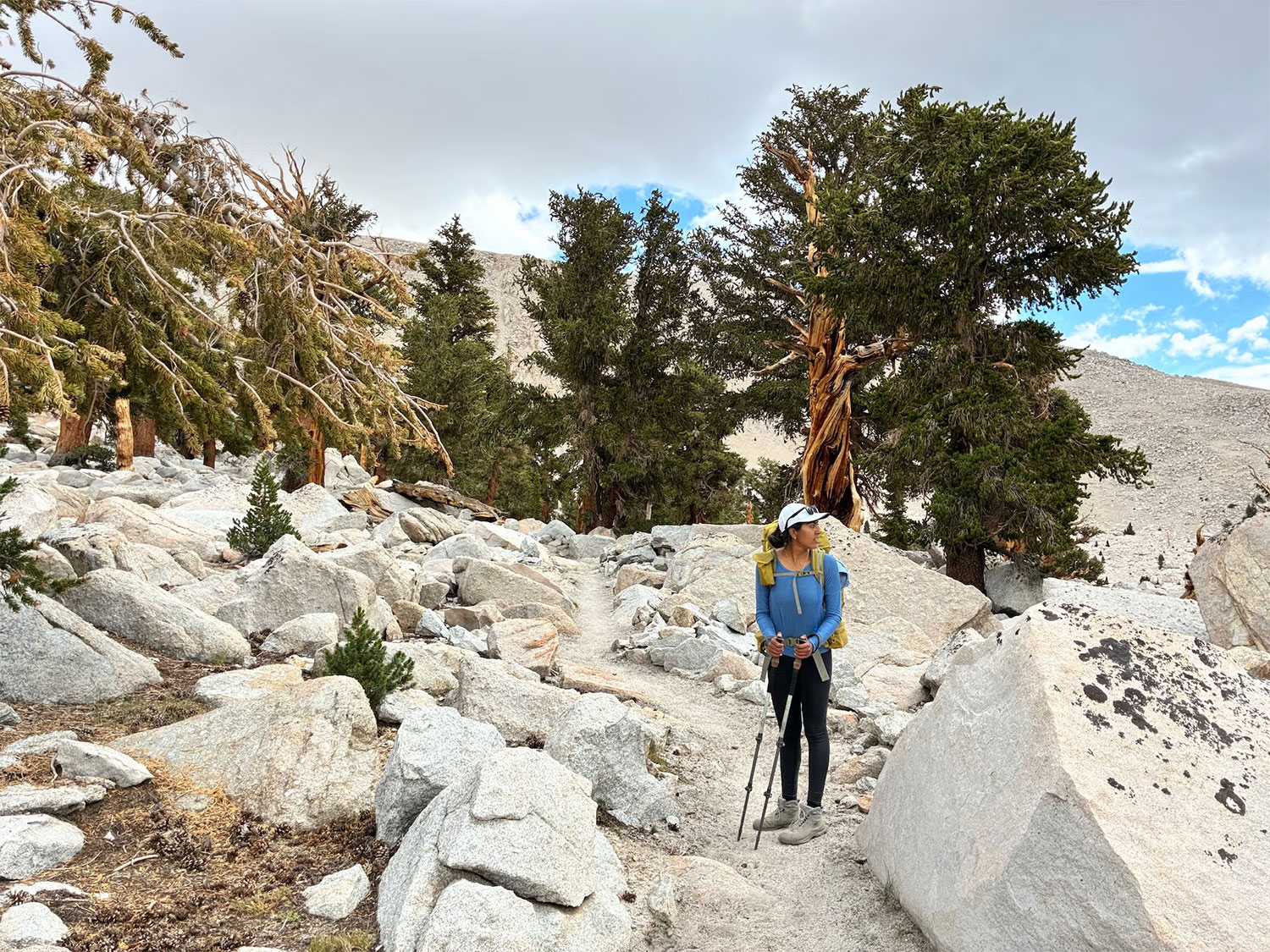 A person in backpacking gear looks out over a mountain vista from a granite trail