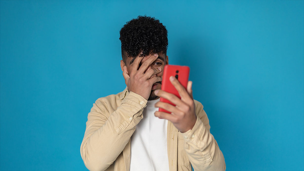 Nervous and scared latino american young man covering his face while reading bad news on the mobile phone against blue background