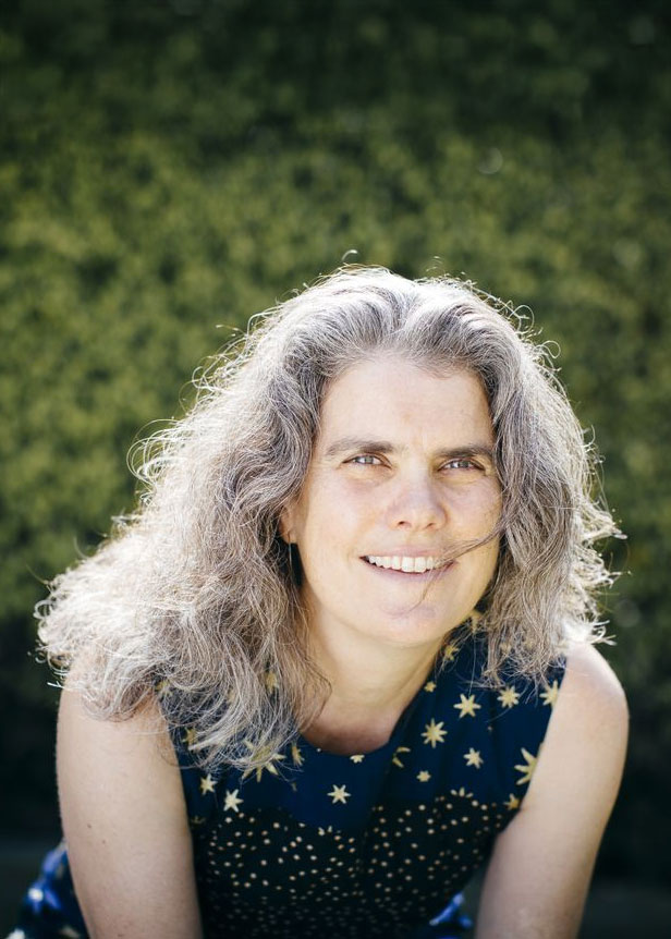 Andrea Ghez smiles at the camera in front of a wall of greenery, wearing a blouse with constellations on it