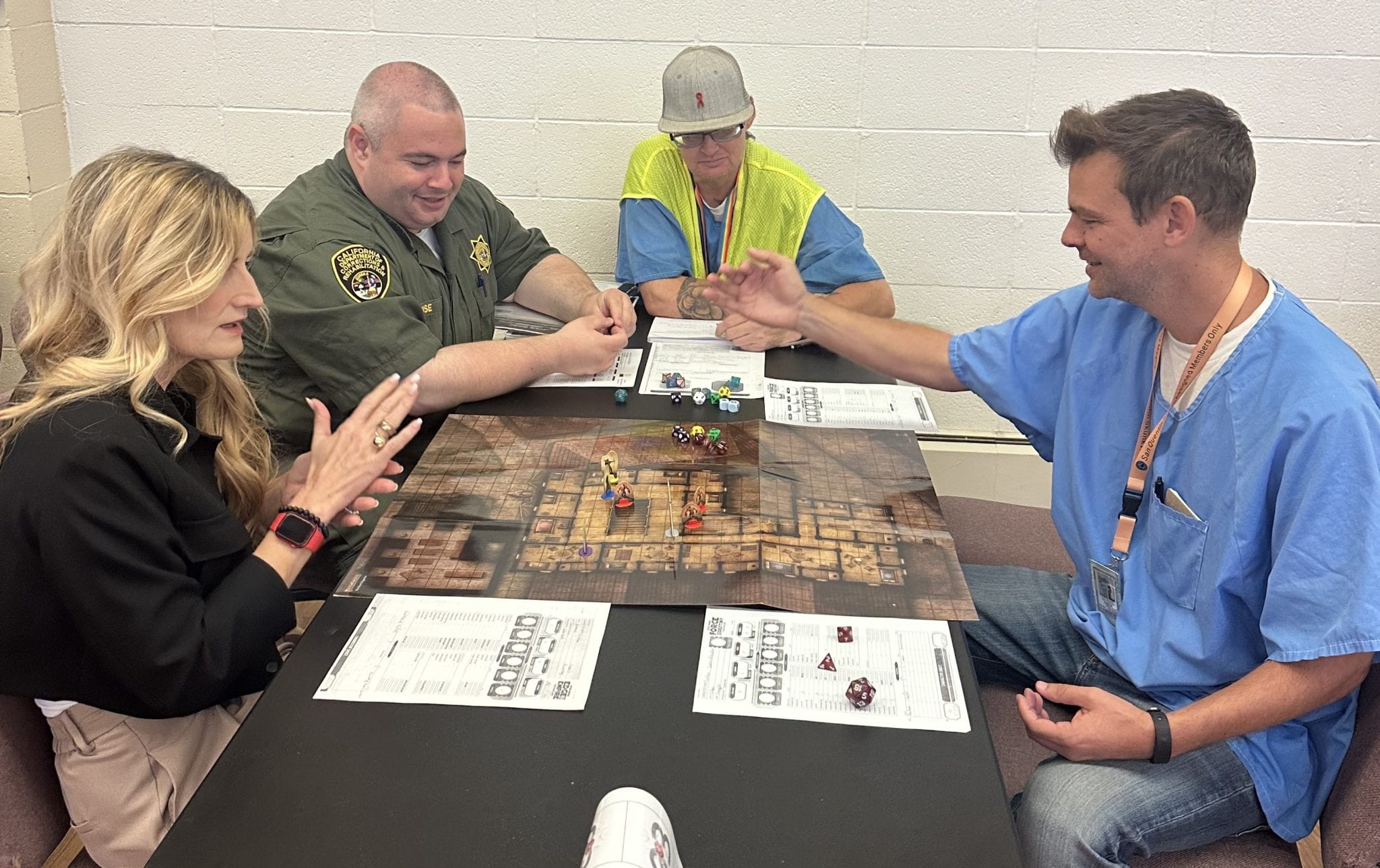 Four people play a tabletop game, a woman on the left, a male sheriff to her left, an incarcerated person on the sheriff's left in a bright vest, and an incarcerated man across from the woman