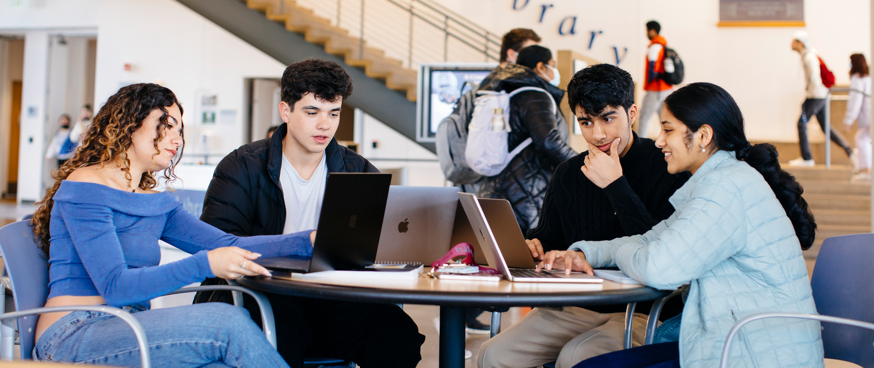 A group of students studying together at a table in a busy campus space, with a sign that says "Library" in the background. 